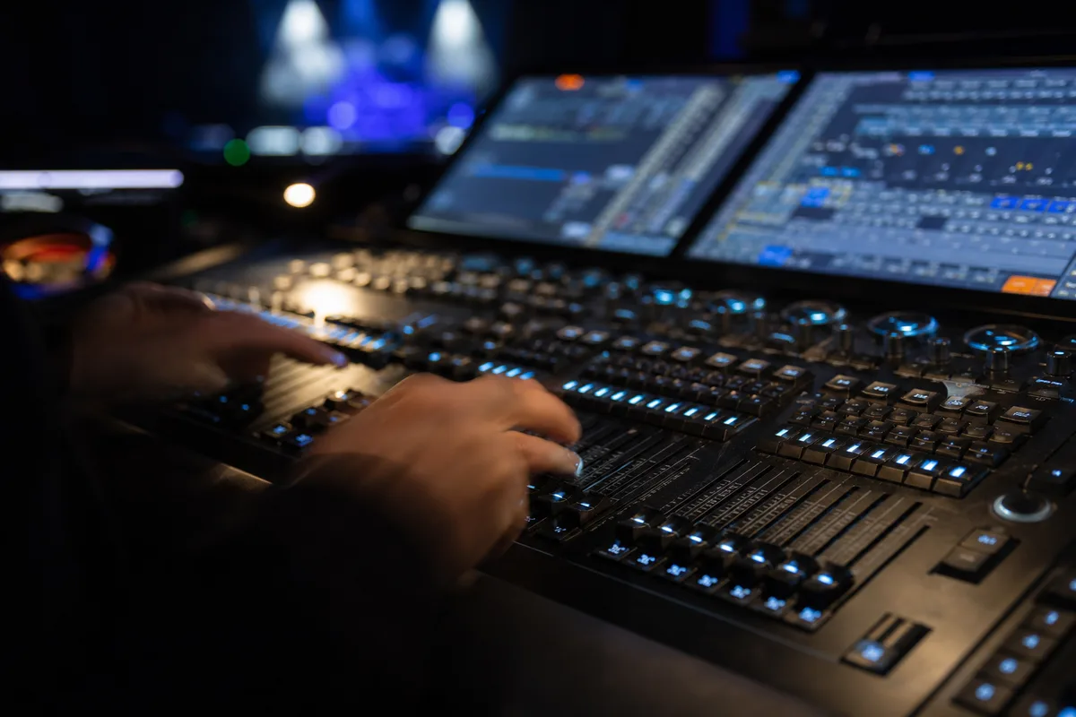 Régie technique d&rsquo;une salle de spectacle, console lumière et faders, écrans de contrôle, mains floues d&rsquo;un technicien, ambiance sombre éclairée par les LED bleutées